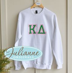 a white sweatshirt with the letters k a on it hanging from a hanger next to a potted plant