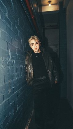 a man standing in front of a brick wall wearing a black sweater and leather jacket