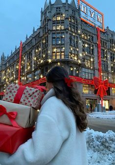 a woman carrying presents in front of a large building with lights on the windows and christmas decorations