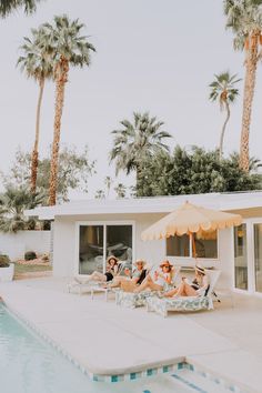 people sitting on lounge chairs by the pool in front of a house with palm trees