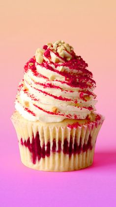 a cupcake with white frosting and red toppings on a plate against a pink background