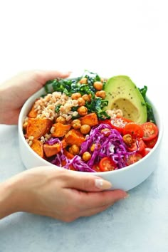 two hands holding a white bowl filled with vegetables and chickpeas, while another hand holds an avocado