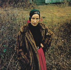 a woman standing in the middle of a field wearing a fur coat and head scarf