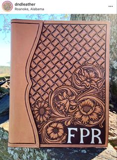 a brown leather journal sitting on top of a tree trunk with flowers and initials in the middle