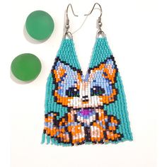 a pair of beaded earrings with an orange and blue cat on it, sitting next to green beads