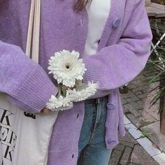 a woman holding a white flower in her right hand and wearing a purple cardigan