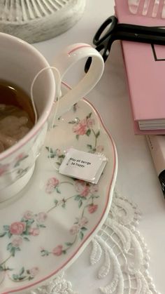 a cup of tea on a saucer next to a pink notebook and computer keyboard