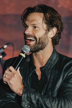 a man holding a microphone in his right hand and smiling at the camera while wearing a leather jacket