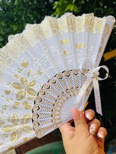 a hand holding a white and gold fan with lace on the top, in front of some trees