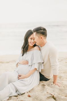 a pregnant couple sitting on the beach in front of the ocean with their belly wrapped around each other