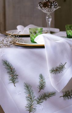 a white table cloth with pine branches on it and two green glasses in the background