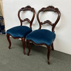 two wooden chairs with blue velvet upholstered seats against a white wall and carpeted floor