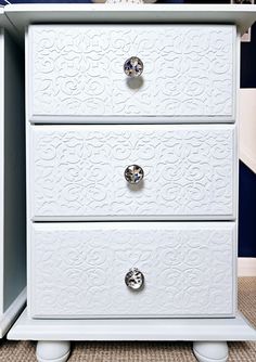 a white dresser with two drawers and knobs on the bottom drawer, in front of a blue wall