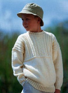 a young boy wearing a white sweater and hat with his hands in his pockets, looking to the side