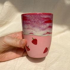 a hand holding a pink and white cup with strawberries painted on the side,