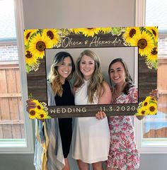 three women standing next to each other in front of a wooden frame with sunflowers on it