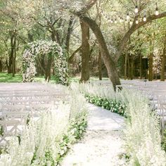 an outdoor ceremony setup with white chairs and flowers on the aisle, surrounded by trees