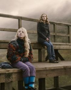 two women sitting on a wooden bench in front of a foggy sky and mountains