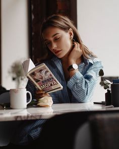 a woman sitting at a table reading a book and holding a coffee cup in front of her