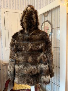 "This unique jacket from Steen and Wright Furriers is made from raccoon fur in natural light to dark brown, with black leather strips between the layers of fur. It has the original leather covered buttons in the front, two front pockets and one inside pockets with the initials \"CAP\", and it's fully lined with black silky lining on top and blue plaid lining on the bottom. The hood is made of fur and leather and is also lined. The measurements, taken with the coat lying flat, are: shoulder to shoulder, 17 inches; armpit to armpit, 20 inches; sleeves, 25 inches; length, 28 inches; bottom edge, 24 inches. Please note that there is some wear on the underside of the front edge where the buttons fasten; otherwise, the coat is in very good condition." Fur And Leather Jacket, Brown Raccoon, Brown Fur Jacket, Fur Coat Short, Short Fur Coat, Raccoon Fur Coat, Unique Jackets, Leather Jacket With Hood, Brown Fur