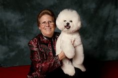 a woman holding a white poodle in her right hand and smiling at the camera