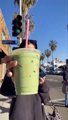 a woman holding up a green drink in front of a traffic light with palm trees behind her