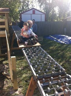 two children are playing in an outdoor play area with metal bars on the ground and wooden structures behind them