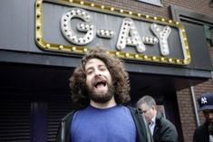 a man with long hair standing in front of a sign that says gay on it