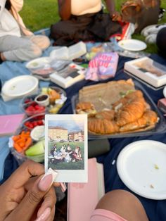 a person holding up a polaroid photo in front of food on a picnic table