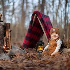 a baby sitting in the woods next to a campfire with a lantern on it