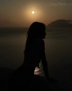 a woman is sitting on the beach watching the sun go down over the water with her long hair blowing in the wind