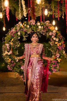 a woman in a pink and gold sari standing under a floral arch at night