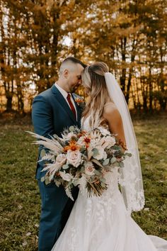 a bride and groom standing together in front of trees with fall foliage on the ground