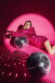 a woman laying on the ground next to two disco balls in front of a pink background