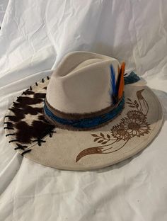 Off white wide brim, Cow horns and flowers burnt on brim. Cowhide material with blue band and feathers. White Brimmed Country Style Felt Hat, White Hat Bands For Spring Ranch, White Western Felt Hat For The Beach, White Hat Band For Spring Ranch Occasions, Spring White Hat Band For Ranch, White Felt Hat With Curved Brim For Rodeo, White Western Felt Hat For Kentucky Derby, White Western Felt Hat For Country Events, White Short Brim Felt Hat For Ranch