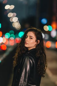 a woman with curly hair standing on the street at night wearing a black leather jacket