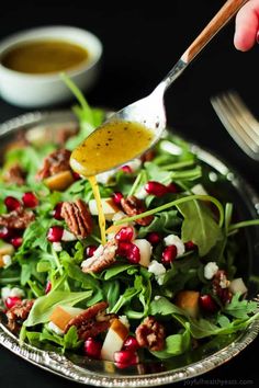 a person pouring dressing onto a salad in a silver bowl with walnuts and pomegranate