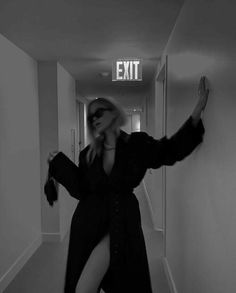 a woman in a long black coat is walking down a hallway with an exit sign above her head