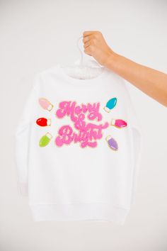 Get your little ones in the festive spirit with our MINI KIDS MERRY & BRIGHT WHITE PULLOVER. This white pullover features a playful pink Merry & Bright patch and festive christmas lights patches. Perfect for adding a touch of holiday cheer to their outfit! All orders are currently shipping within 14 business days. To receive item quicker, expedited shipping is available at checkout. **All Christmas orders must be placed with expedited shipping to guarantee delivery by Dec. 24 if placed after Dec Festive Pink Crew Neck Top, Cute White Christmas Sweater, Pink Cotton Holiday Sweatshirt, Pink Holiday Festive Tops, Pink Holiday Festive Top, Pink Holiday Tops For Festive Occasion, White Festive Holiday Sweater, White Holiday Festive Sweater, Fun White Sweatshirt For Winter