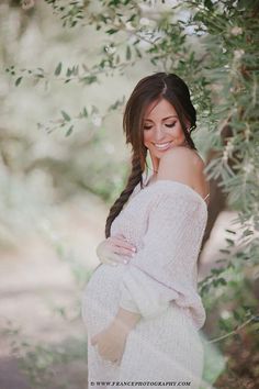 a pregnant woman is smiling while standing under an olive tree with her hands on her stomach