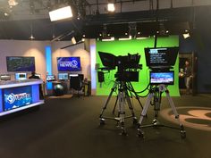 three cameras are set up on tripods in front of green screen television studio monitors