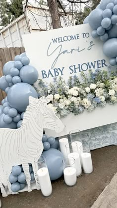 a baby shower sign with blue balloons and white flowers in front of it, surrounded by candles