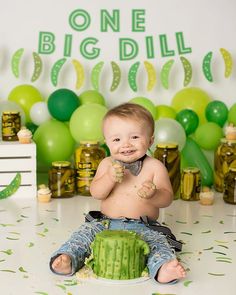 a baby boy sitting on the floor in front of a cake with green balloons and confetti