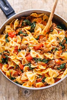 a skillet filled with pasta and spinach on top of a wooden table next to a spatula