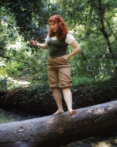 a woman with red hair standing on a log in the woods, holding a stick