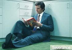 a man in a suit sitting on the floor reading a book with his legs crossed