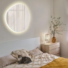 a cat laying on a bed in front of a round mirror with lights above it