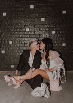 a man and woman sitting next to each other in front of a brick wall kissing