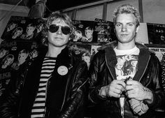 two men standing next to each other in front of a wall with posters on it