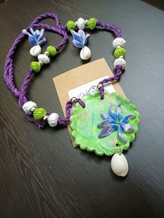 a purple and green necklace with flowers on it sitting on a table next to a tag